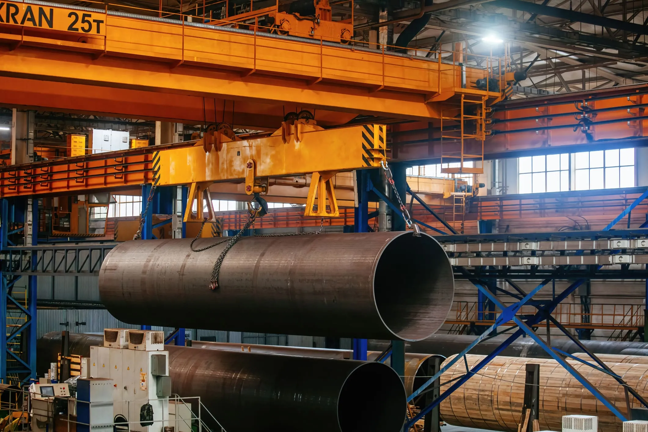 Overhead crane moving large steel pipe through an industrial fabrication plant